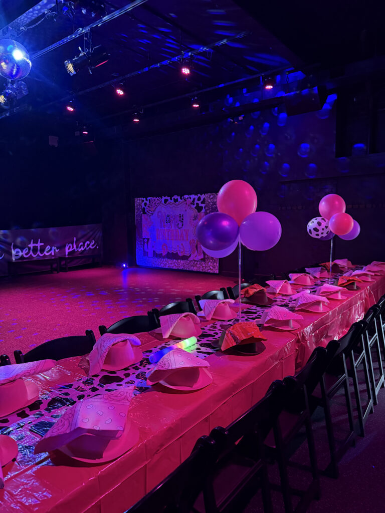 Party room with pink and blue lighting, long table with cowboy hats, birthday banner, and balloons.