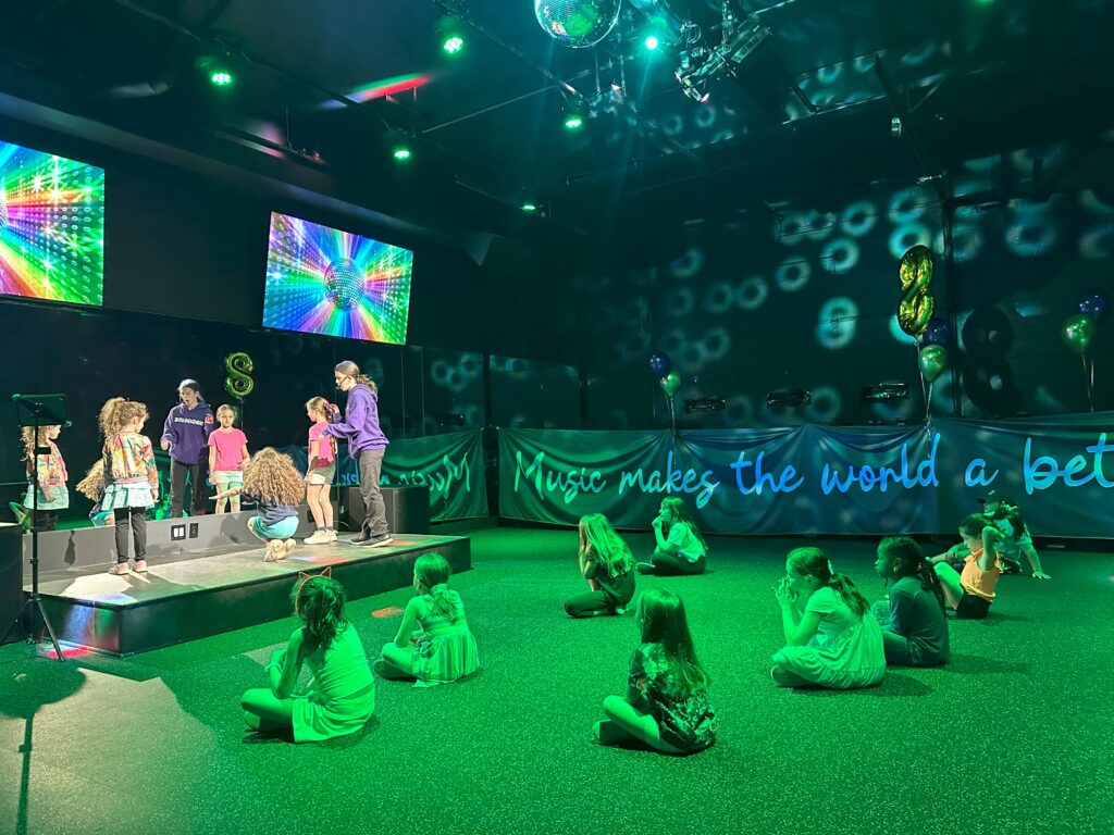 Children performing on stage with colorful lights and screens, celebrating music and creativity in a vibrant setting.